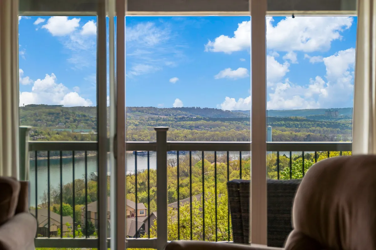 The image showcases a sweeping, panoramic view of Table Rock Lake from the condo, framed by rolling, lush green hills and a vast blue sky dotted with clouds. The scene exudes serenity, with sunrise or sunset hues adding warmth and inviting moments of peaceful relaxation. The vantage point suggests guests can enjoy the view from indoors or step out onto a balcony.