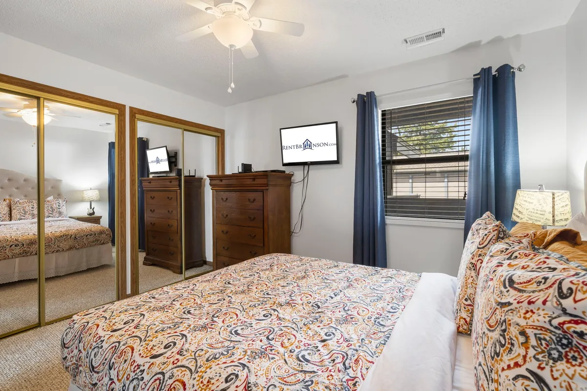 A cozy secondary bedroom with a comfortable bed dressed in colorful bedding, a mirrored wardrobe, wooden dresser, and a wall-mounted television. The room feels welcoming and practical, ideal for guests or family members to unwind after a busy day.
