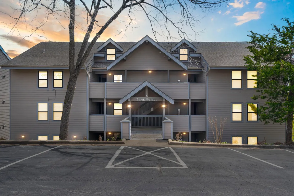 This image captures the exterior entrance of the condo building, framed by large windows and a leafy tree in the foreground. The scene conveys both accessibility and a welcoming atmosphere, with a glimpse of the surrounding environment that hints at proximity to local attractions.