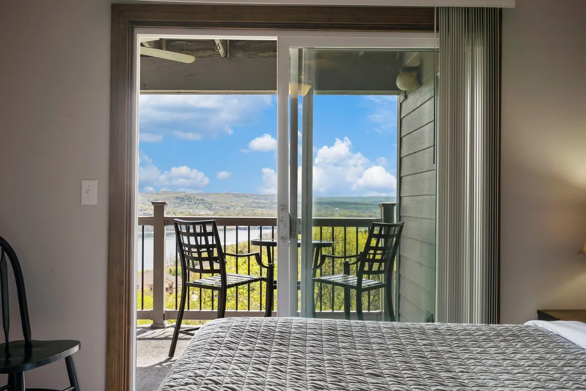 A cozy bedroom with a bed facing sliding glass doors that open onto the deck, showcasing chairs outside and a sweeping lake view. Natural light floods the room, and the direct access to the balcony highlights the seamless indoor-outdoor living experience.