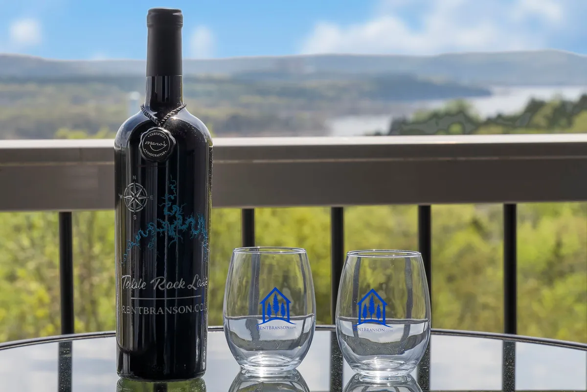 A bottle of wine and two glasses set on a balcony table, overlooking a scenic, sunlit expanse. The arrangement suggests an intimate, relaxing atmosphere for watching sunsets or enjoying a quiet drink while taking in the lake view.