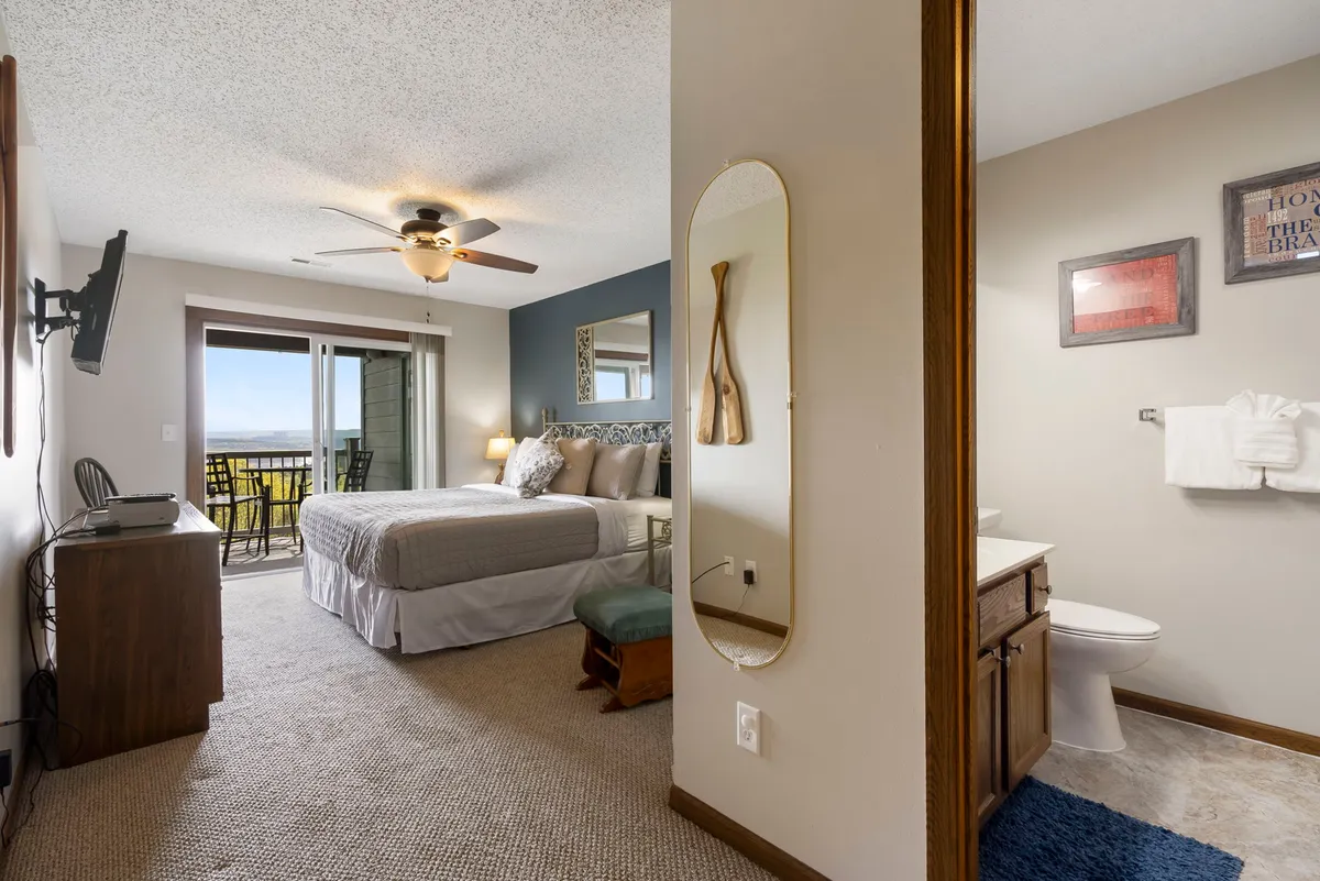 A serene master bedroom featuring a plush bed adorned with decorative pillows, a sliding glass door opening to a private balcony, and a well-lit vanity area with a mirror. The room feels tranquil and inviting, with soft lighting and calming colors, hinting at restful nights and easy mornings.
