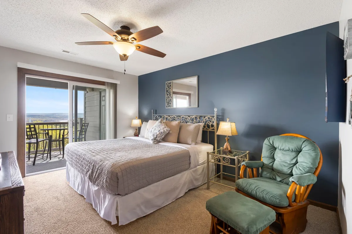 A serene bedroom with a plush bed, an armchair, and large balcony doors opening to the outside, inviting natural light. The scene hints at private mornings with coffee while enjoying the lakeside view.