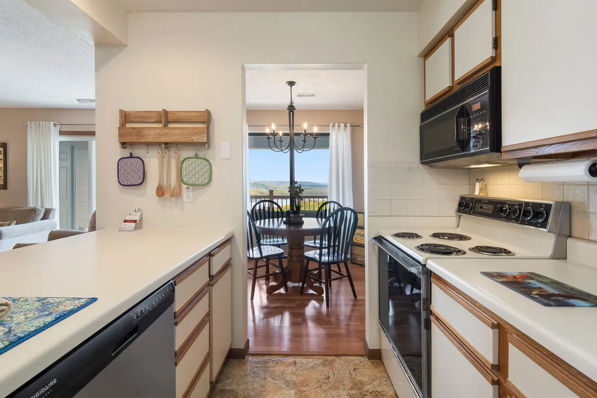 A warm kitchen and dining area is pictured, blending black appliances, a kitchen countertop, and a dining table set beneath a chandelier. The view through the doorway hints at further open space and windows, suggesting the inviting continuity of the home.