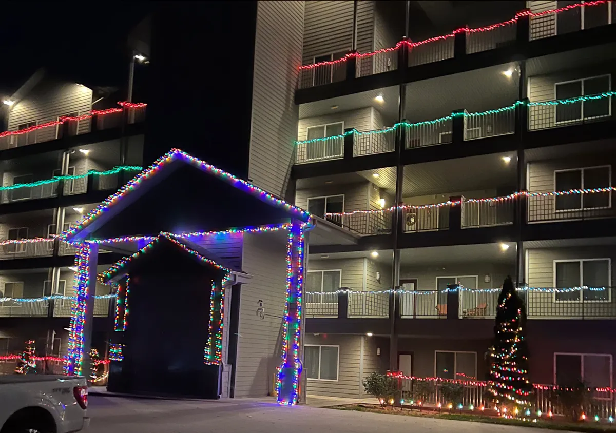 A decorated building with sparkling holiday lights, an illuminated entrance, and a Christmas tree sets a festive scene. The mood is joyful and inviting, suggesting community events and gatherings during the holiday season.